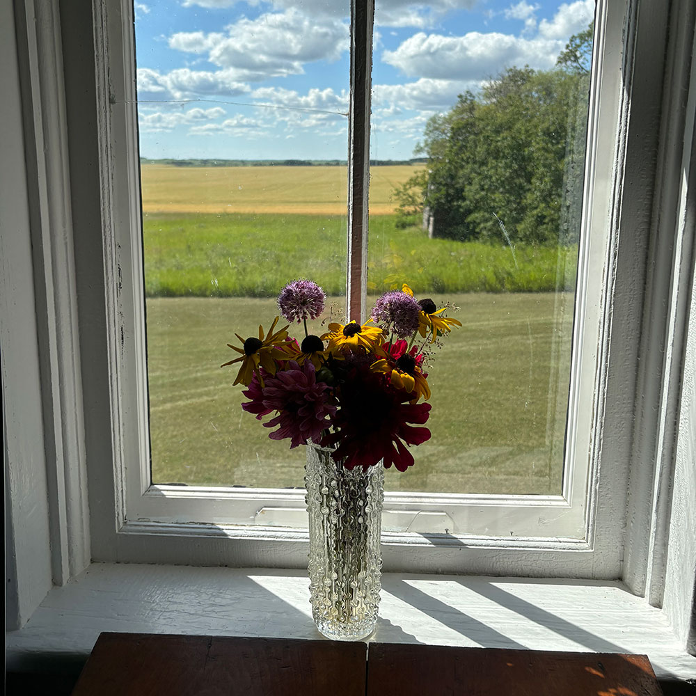 A bouquet of red, orange and purple wildflowers sits on the sill of the west window of Kingsley School on August 10, 2024. Through the window can be seen a prairie vista of grassland and fields and a blue, partly cloudy sky. The day is bright and sunny.
